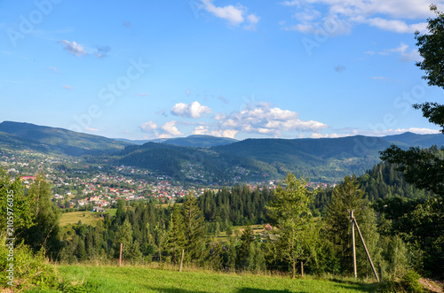 Konstfotografi Panoramic view of a mountain village nestled in a green valley, surrounded by forests and rolling hills under a bright blue sky with fluffy clouds