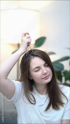A relaxed brunette woman uses a handheld scalp massager on her head, eyes shut in pure bliss and wellness, capturing peaceful self-care moments indoors near plants.