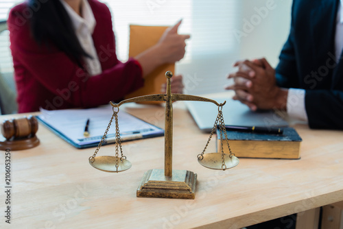Close up of justice scales and judge gavel on wooden desk with cash bundle and lawyer consulting client about financial lawsuit in blurred background.