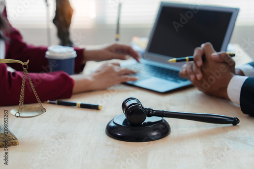 Close up of justice scales and judge gavel on wooden desk with cash bundle and lawyer consulting client about financial lawsuit in blurred background.