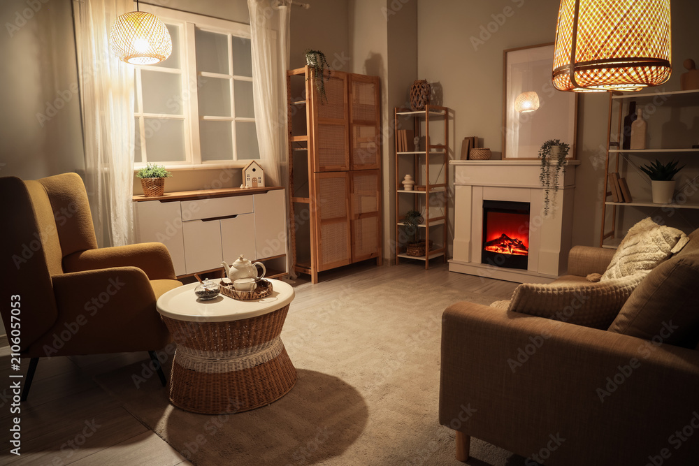 Obraz premium Cozy living room interior with warm lighting, comfortable armchairs, and a decorative electric fireplace at night