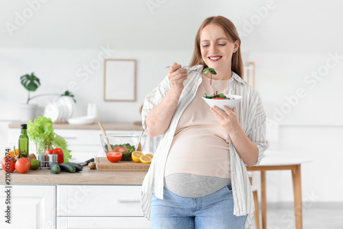 Young pregnant woman eating vegetable salad in kitchen