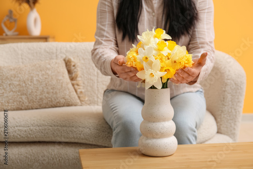 Woman sitting near vase with narcissus flowers and touching them in room