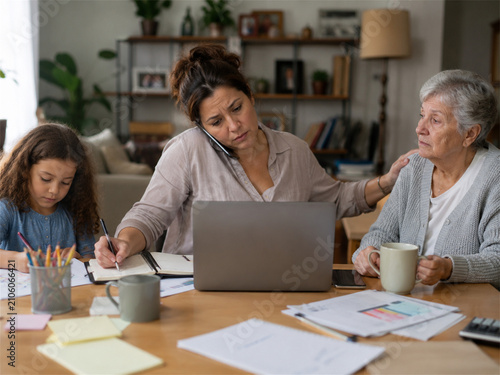 Overwhelmed mother balances remote work and family caregiving at a kitchen table while supporting an elderly woman and supervising a child during a demanding day at home and household responsibilities