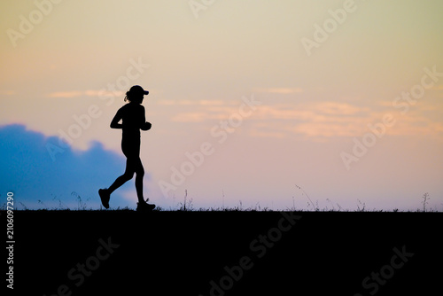 Quadro su tela Side view silhouette of a female runner jogging at sunset along a scenic horizon with warm orange tones and soft blue sky showcasing fitness endurance active lifestyle and motivation