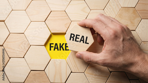 Hand placing wooden hexagon tile reading real over fake text