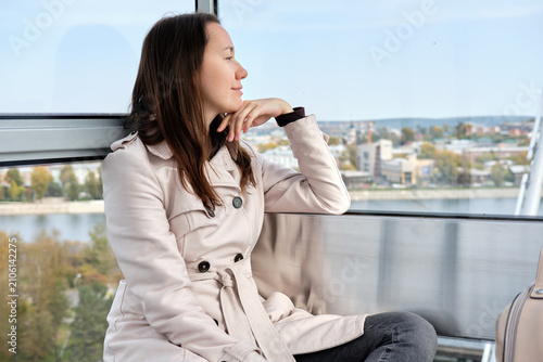 Fotografie A young woman in a beige trench coat sits inside a modern ferris wheel cabin, quietly admiring the city skyline through a panoramic window