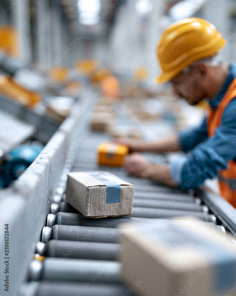 Fototapeta premium Worker in hard hat handles packages on conveyor belt. Focused on task, eyes down, hands steady. Boxes move steadily past with mechanical precision. Ideal for logistics, warehousing