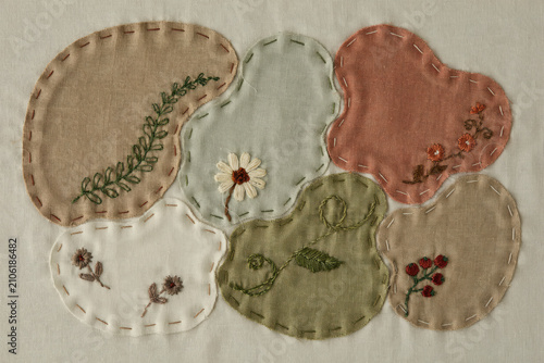 Hand embroidered fabric patches in irregular organic shapes featuring floral and leaf motifs stitched onto a plain white linen background