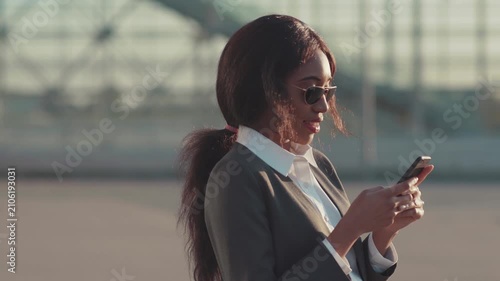Beautiful long-haired Afro-American woman in trendy sunglasses actively uses her smartphone outside the office, gets good news, looks happy and satisfied. Positive emotions, cheerful mood, feeling