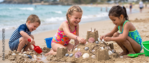 Three children building a sandcastle on a sandy beach. Happy kids playing with toys and seashells by the ocean during summer vacation