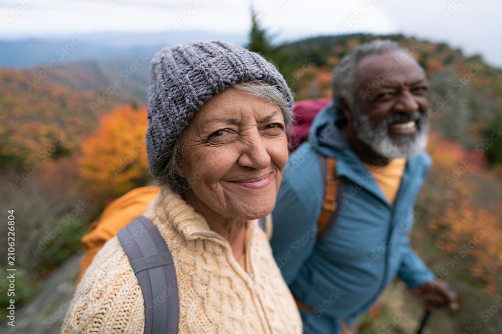 Fototapeta premium Older couple smiles warmly on autumn trail. Backpacks hint at a scenic hike. Mountain range stretches behind them. Vibrant fall colors surround the path. Their joy reflects shared adventure