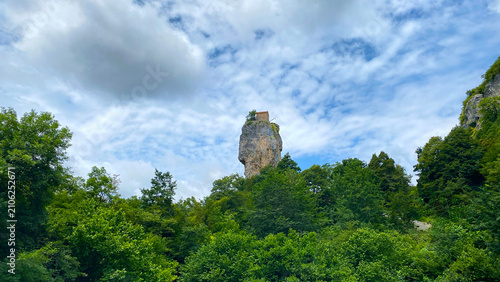 Katskhi Pillar stands isolated above a forest in a distant wide view, with the hilltop church framed by layered clouds in Imereti