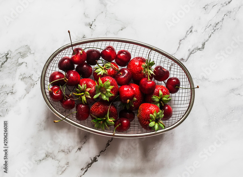 Fresh strawberries and cherries in a metal basket on a light marble background, top view
