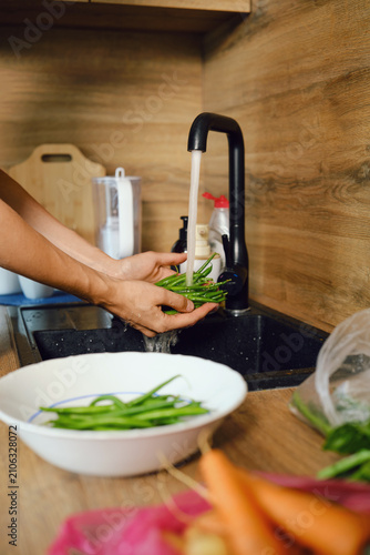 Hands hold green beans under running water in kitchen sink before meal preparation