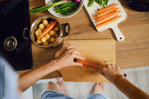 Top view of hands slicing fresh carrot lengthwise on wooden chopping board in kitchen
