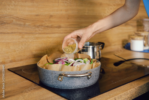 Person sprinkles spice and herbs to roasting pot with vegetables in kitchen