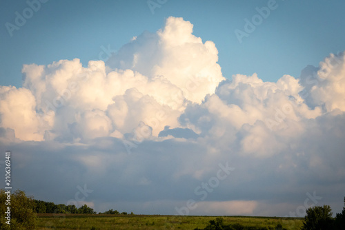 Vast cumulus towers rise over a sunlit meadow and distant treeline while warm evening tones wash the landscape evoking the stillness before summer storms on a peaceful countryside afternoon