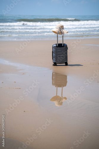 A straw hat resting on top of a grey rolling suitcase on a sandy beach with the sea behind and a mirror-like reflection of both hat and suitcase in the wet sand below. Vacation awaiting concept