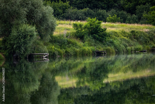 Konstfotografi A lush riverbank of willows and reeds reflects in quiet water with old fishing p
