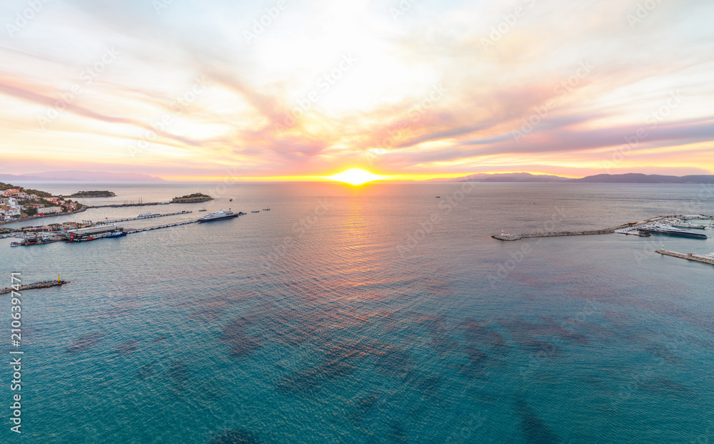 Obraz premium Kusadasi, Turkey. Aerial view of blue bay with Pigeon Island, port, and distant mountains. Sun at horizon with smoky sky. Aerial view.
