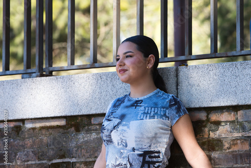 A young, pretty Gypsy woman from Seville, with long dark hair tied back in a ponytail and dressed in casual clothes, is leaning against a wall, smiling happily.