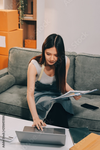 фотографія Startup SME small business entrepreneur of freelance Asian woman wearing apron using laptop and box to receive and review orders online to prepare to pack sell to customers, online sme business ideas