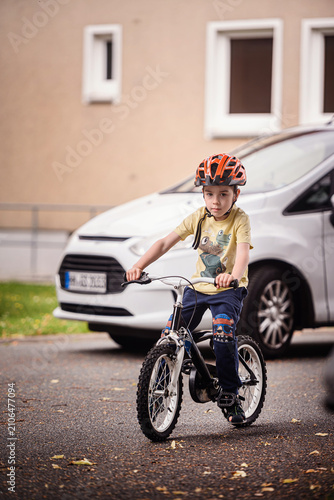 Preschooler boy on bicycle with orange safety helmet riding past parked white car on suburban street
