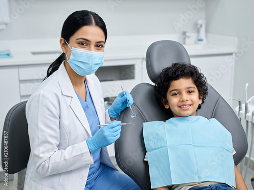 young fair skinned indian female dentist examining smiling young boy during routine pediatric dental checkup in modern clinic promoting oral health
