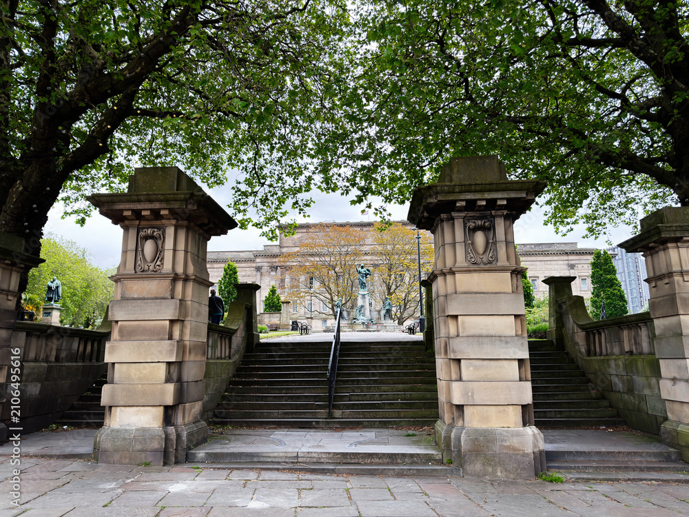 Fototapeta premium Grand stone St. John's Gardens entrance stairs and neoclassical museum facade framed by leafy trees, ornamental pillars and statues in Liverpool, UK