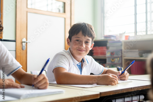Tableau sur toile Smiling schoolboy writing in notebook during classroom lesson