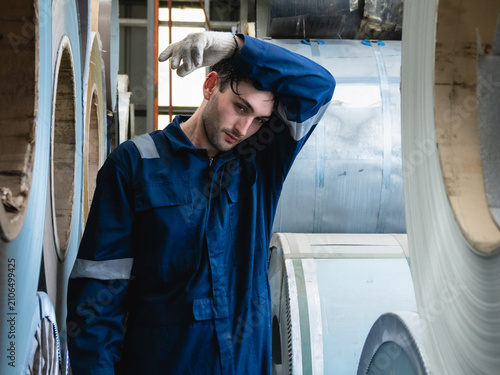 close-up portrait of an exhausted male industrial worker in blue overall wiping sweat from his forehead with his glove forearm while working among heavy steel coil inside warehouse.
