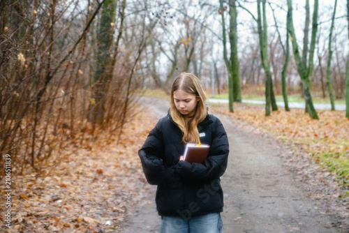 Thoughtful Young Woman Holding Book Walking on Autumn Park Path in Melancholy Mood