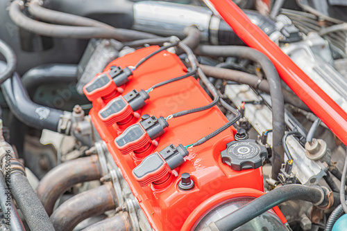 Car engine close-up featuring red distributor cap, ignition coils, bundled wires, metallic hoses, rubber tubes, well-maintained condition, clean appearance, natural lighting