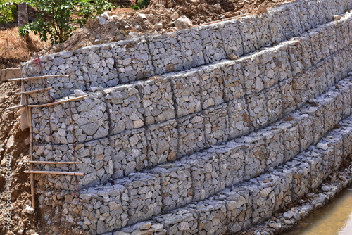 Retaining wall constructed from gabion baskets filled with stones viewed from the side outdoors, Gabion retaining wall