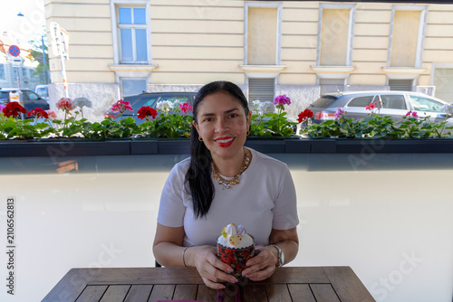 A smiling woman sitting at a wooden table in a cafe, holding a small decorated dessert cup with flowers blooming in a window box behind her.