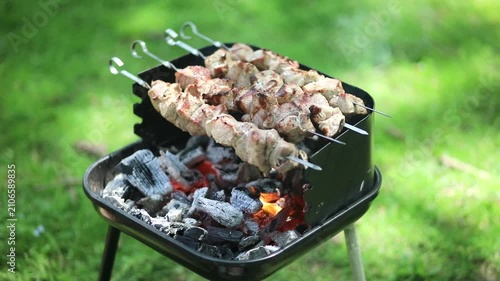 Meat skewers grilling over hot charcoal on a barbecue grill outdoors, with flames, smoke and green grass in the background