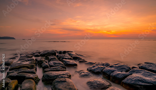 Foto Long exposure sunset seascape with rugged coastal rocks, silky ocean water and glowing orange twilight sky, creating a peaceful tropical shoreline landscape