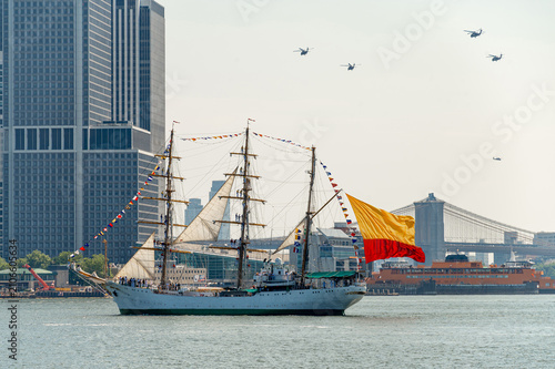 Tall ship carries Colombian flag beneath helicopter formation. Sailing vessel passes Lower Manhattan with Brooklyn Bridge, ferry terminal and aircraft in the background.