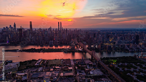 New York City skyline at sunset, view of Manhattan and Queensboro Bridge. Aerial view of the East River and skyscrapers under an orange sky.