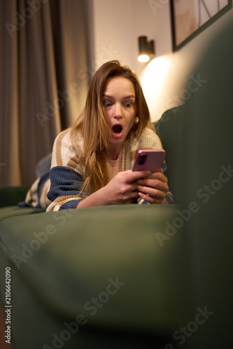 Woman lying on green sofa looking surprised at phone screen