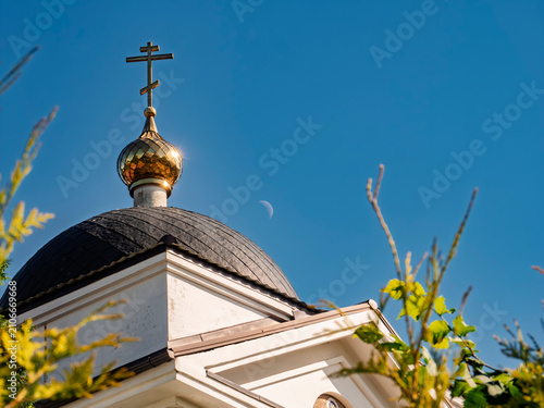 Golden cross and dome of an Orthodox church against a clear blue sky with a crescent moon