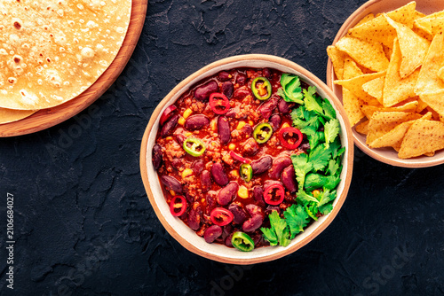Spicy chili con carne, traditional Mexican beef stew. Flat lay on black slate background with tortillas, nachos