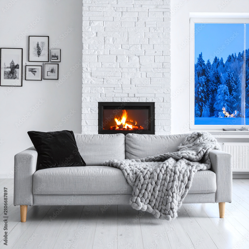 Obraz premium A cozy living room interior featuring a modern grey sofa, warm fireplace, knit blanket, and snowy winter forest window view, evoking warm comfort and serene peace. There are no people.