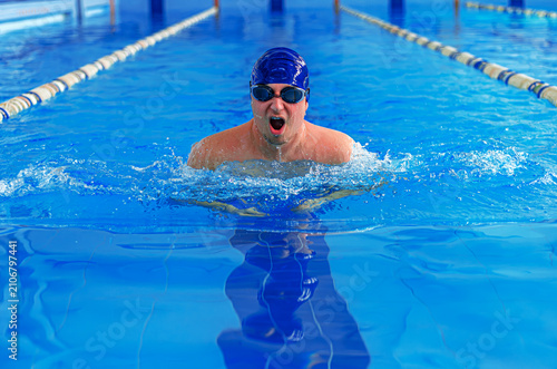 A male swimmer wearing a blue swimming cap and goggles takes a breath while swimming in a pool.