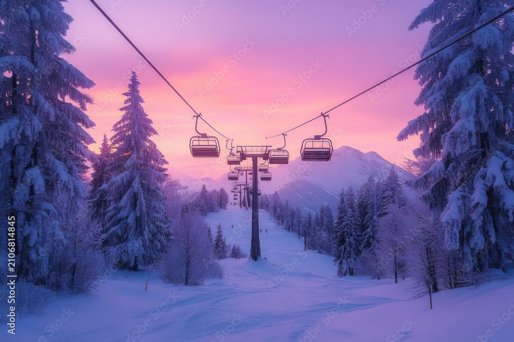 Obraz premium empty ski chairlift gliding above a snow-covered trail flanked by frosted pine trees toward a distant mountain under a pastel pink and purple sunrise, tranquil and magical