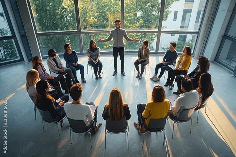 Obraz premium diverse group of adults seated in a circle indoors with a standing facilitator near large windows, warm sunlight and an engaged, open collaborative atmosphere