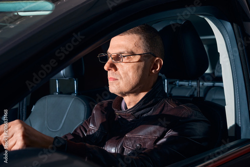 Serious mature man in glasses driving a car at night, side view through the window