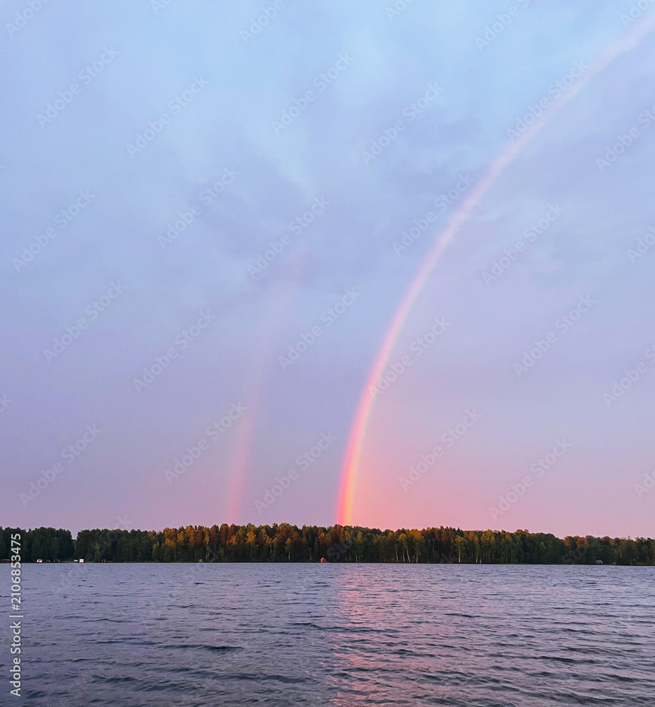 Obraz premium Double rainbow over calm lake after rain with forest on the horizon