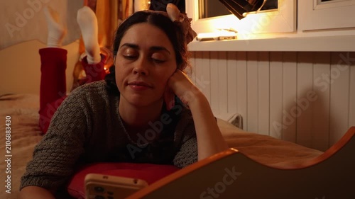 Beautiful young Caucasian woman smiling while using a smartphone lying on a bed at home in the evening. Social media, online communication, digital lifestyle, and cozy home relaxation concept.